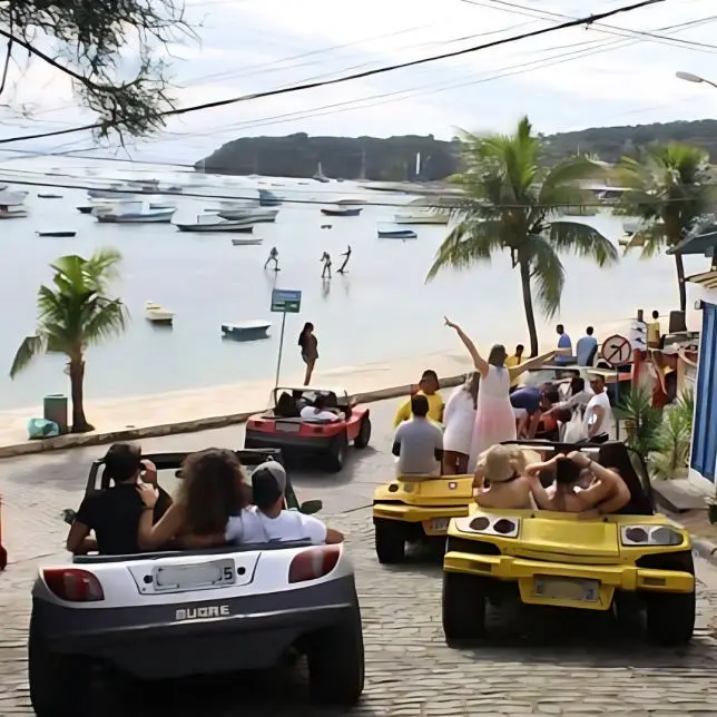 Fila de buggies descendo uma ladeira de pedra rumo a uma enseada de Búzios, com barcos ancorados e banhistas na praia ao fundo.