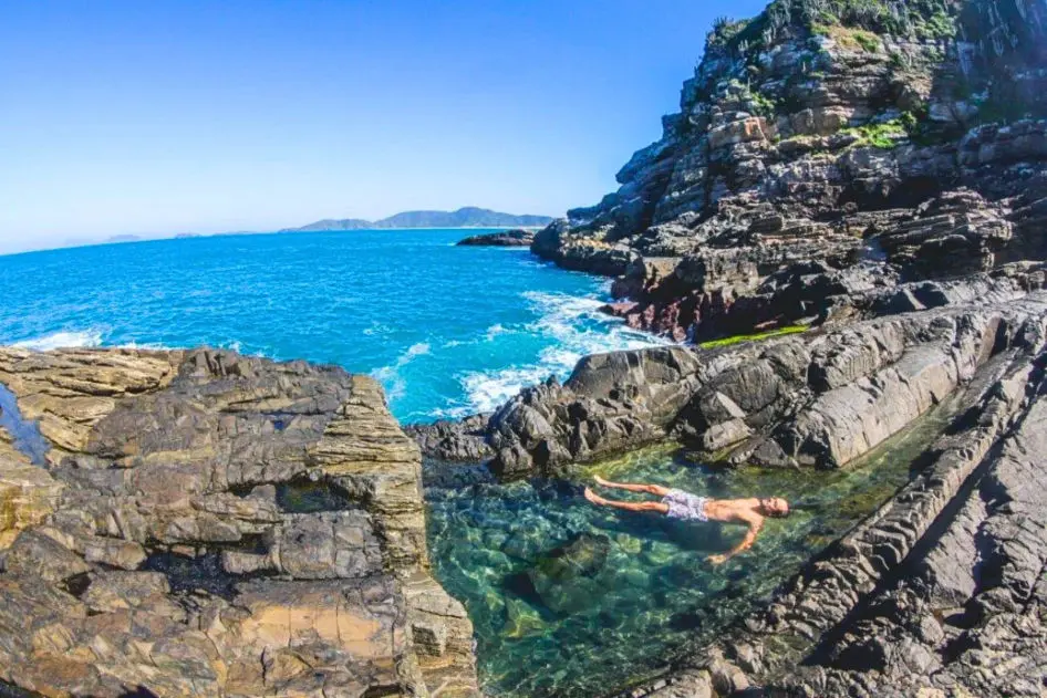 Pessoa flutua em piscina natural de água cristalina cercada por formações rochosas numa trilha de ecoturismo em Búzios