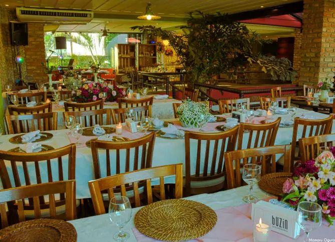 Salão do restaurante Buzin preparado para um evento privado, com mesas postas, arranjos de flores e iluminação de fim de tarde.
