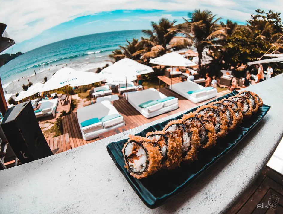 Prato de sushi servido em restaurante de praia com espreguiçadeiras e mar ao fundo em Búzios