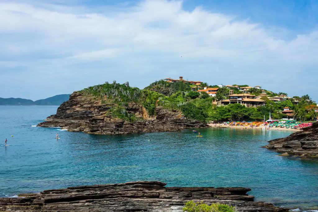Vista aérea da praia de Ferradurinha, em Búzios, com costão rochoso, casas na parte alta e pessoas em pé sobre pranchas de stand up paddle.