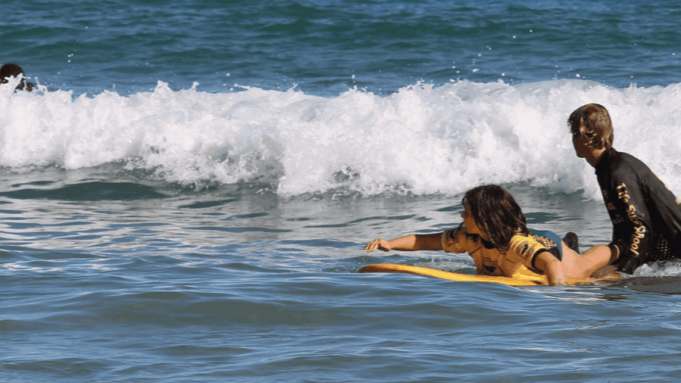 Instrutor auxiliando aluna a pegar uma onda durante aula de surf na Praia de Geribá
